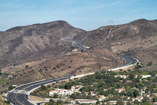 Curving highway running through dry brown hills with scattered homes and power lines under a clear blue sky.